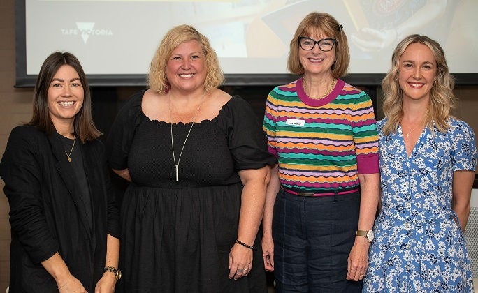 Smiling nursing student stands in training ward, with fellow students in background working on text equipment.