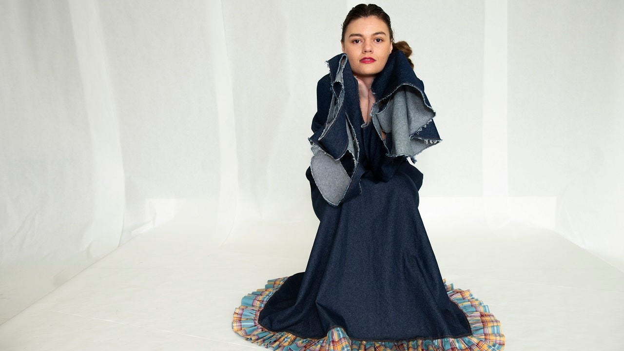 Fashion student in dark blue dress poses in front of plain white background.