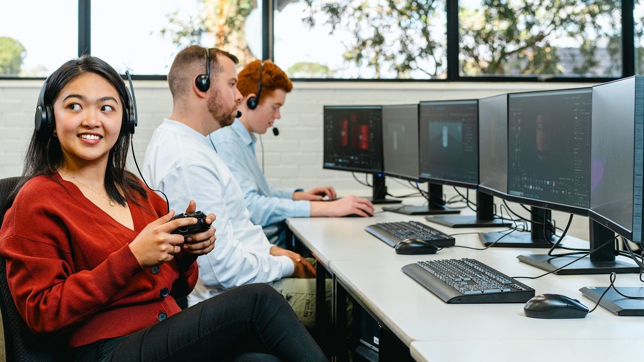 Three students sit in a computer lab, with headsets.