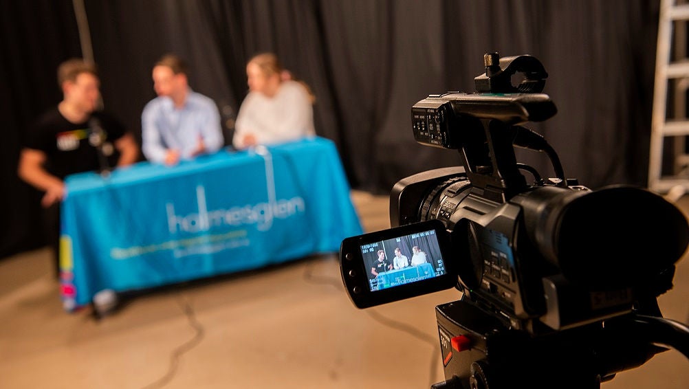 Three students sit in front of a camera for a student broadcast.