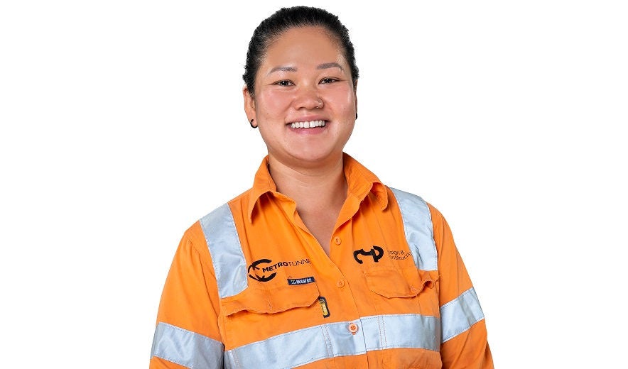 Female civil construction apprentice, wearing orange hi-ves, smiles to the camera.