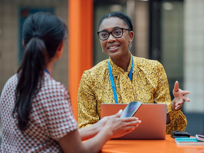 Two female teachers sitting at a table, having a discussion in the lobby area in the school they work at in Gateshead, North East England. They are talking about school issues together while using a laptop.