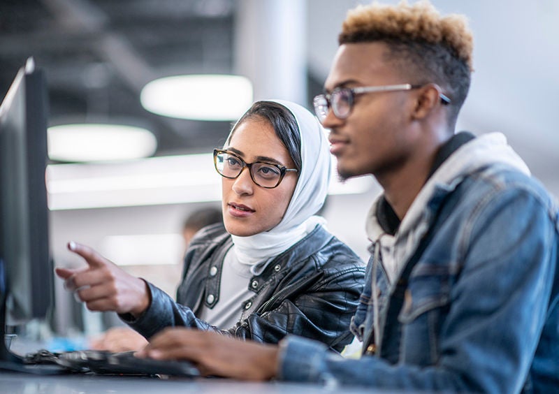 A young man and woman are sitting in a computer lab. The woman is helping with a computer project.