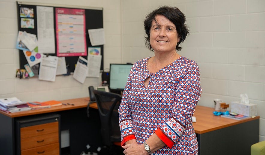 Female teacher stands in office, with desk in background.