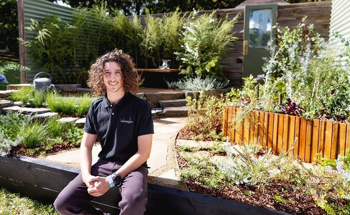 Landscape construction course student sits in front of garden on a sunny day.