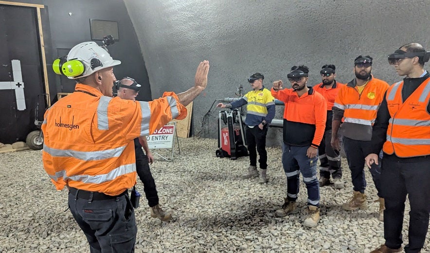 A teacher, in orange shirt and hard hat, teaches apprentices in a tunnelling workshop.