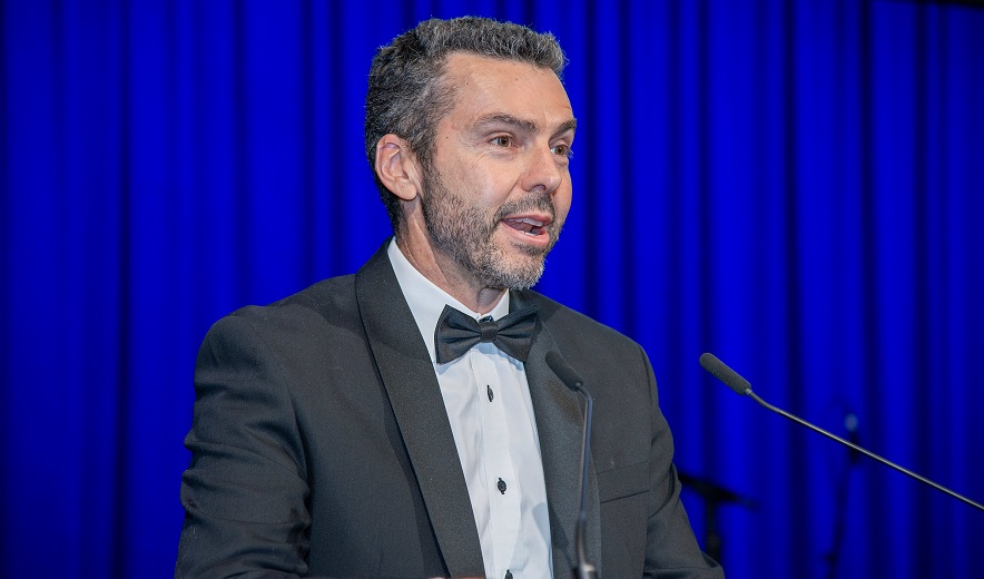 A man in black tie and suit gives speech into microphone at Awards ceremony.