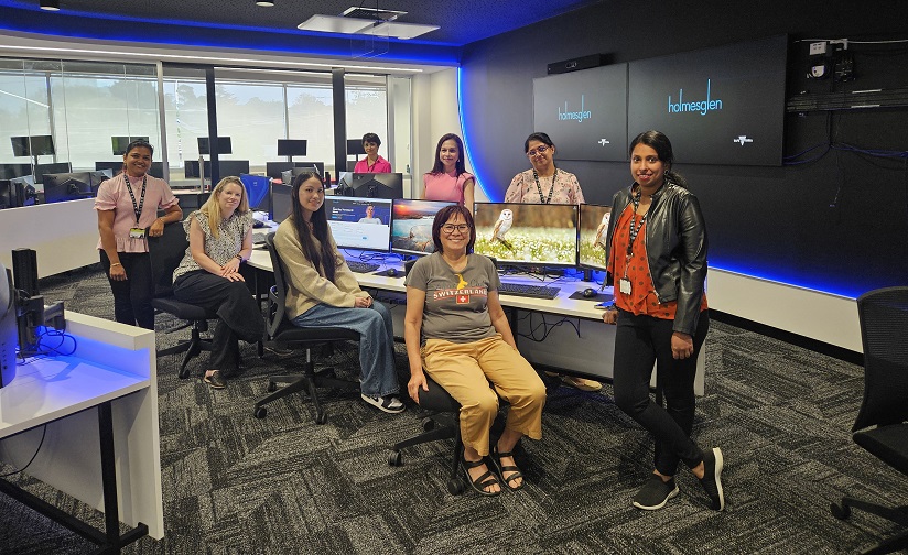 A group, composed of eight IT students and teachers, stand together in Holmesglen's Cyber Security Operations Centre.