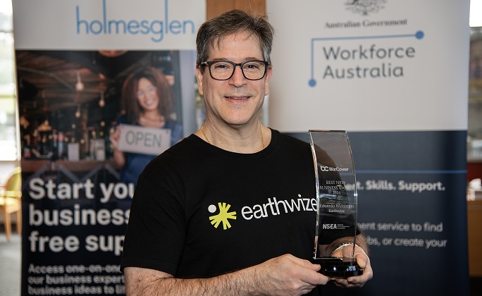 A man in a black t-shirt smiles to the camera, while holding an award trophy.