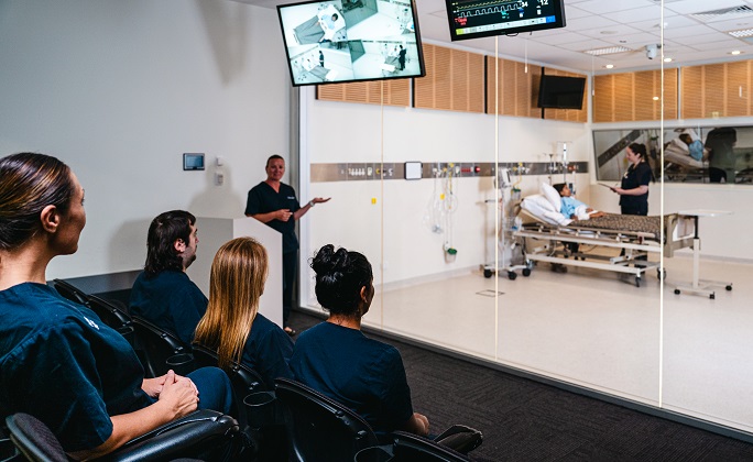 A group of nursing students listen to their teacher, while watching a simulated ward exercise.