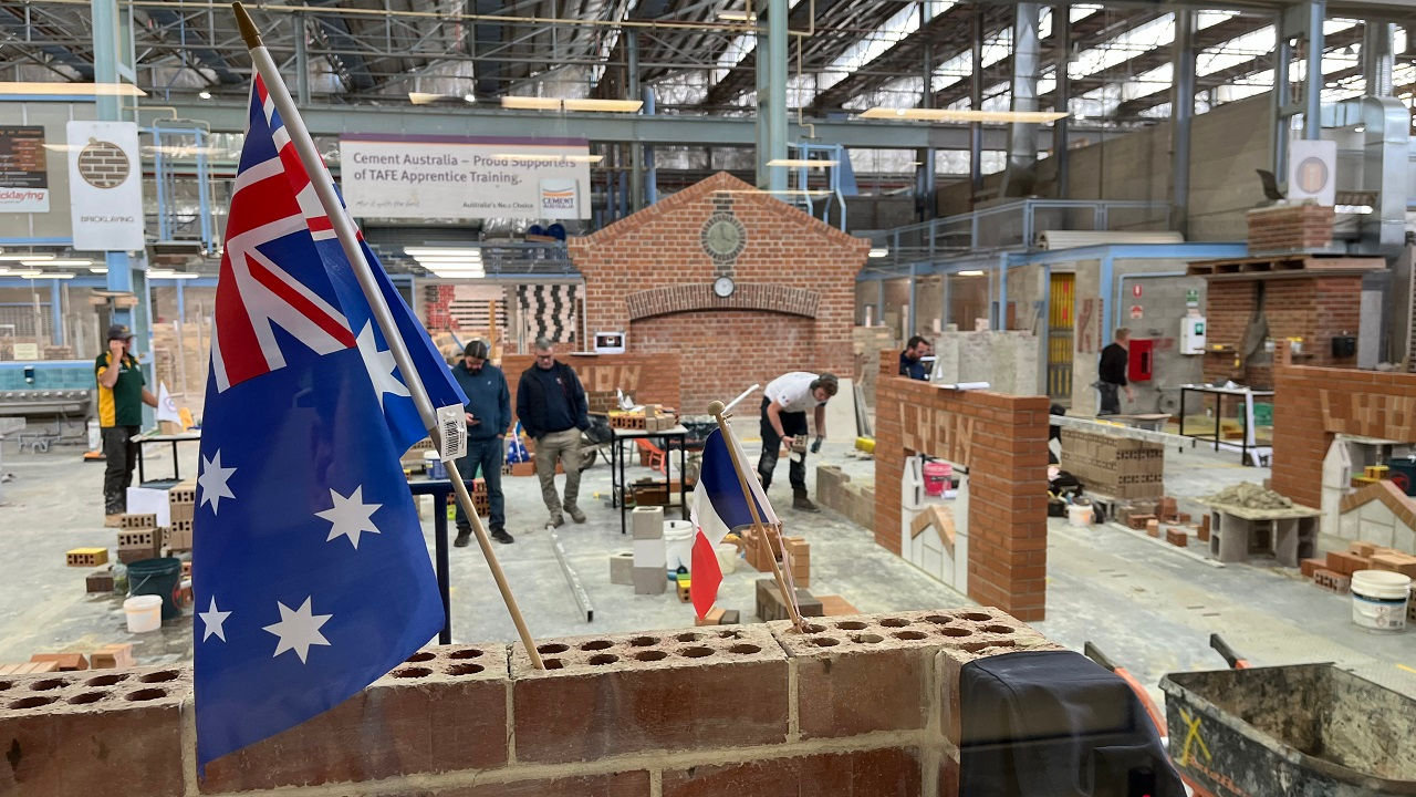 Bricklaying apprentices work on a project in a trades workshop. The Australian and French flags are in the foreground.