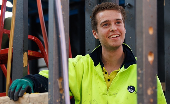 Male plumbing apprentice wearing a hi-vis jumper, smiles while working on an installation in a training workshop.