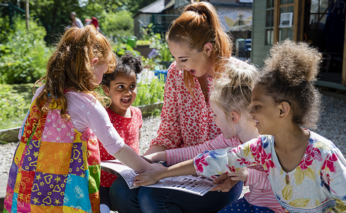 A front view of a teacher and her excited little pupils sitting and doing storytime outdoors in the schoolyard on the benches on a wonderful summer's day in Hexham in the North East of England.