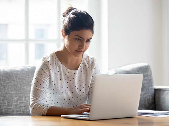 Focused Indian woman using laptop at home, looking at screen, chatting, reading or writing email, sitting on couch, serious female student doing homework, working on research project online