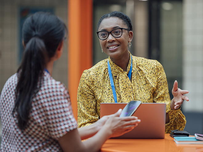 Two female teachers sitting at a table, having a discussion in the lobby area in the school they work at in Gateshead, North East England. They are talking about school issues together while using a laptop.