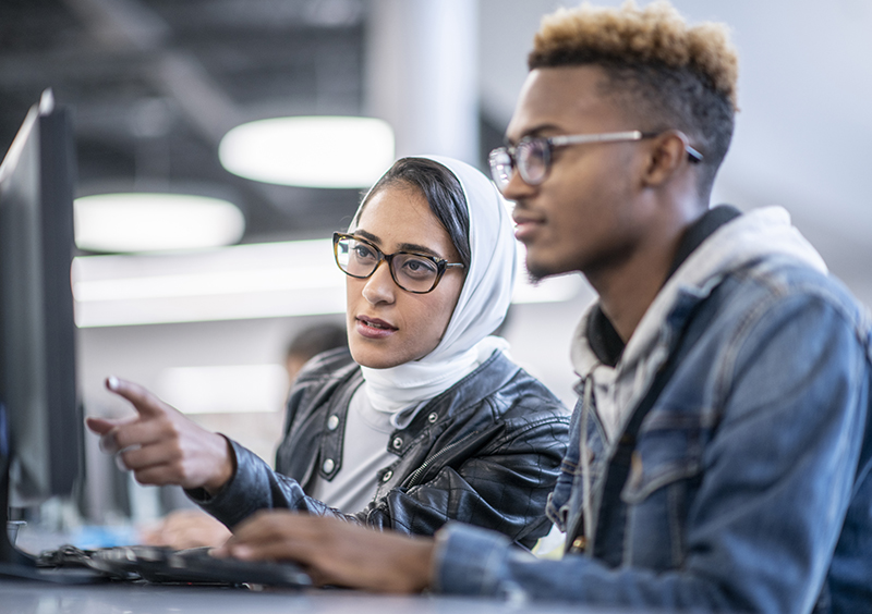 A young man and woman are sitting in a computer lab. The woman is helping with a computer project.