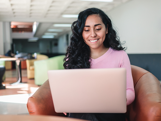 Young hispanic student woman using a computer to study, sitting on an armchair in college campus corridor. Back to school concept