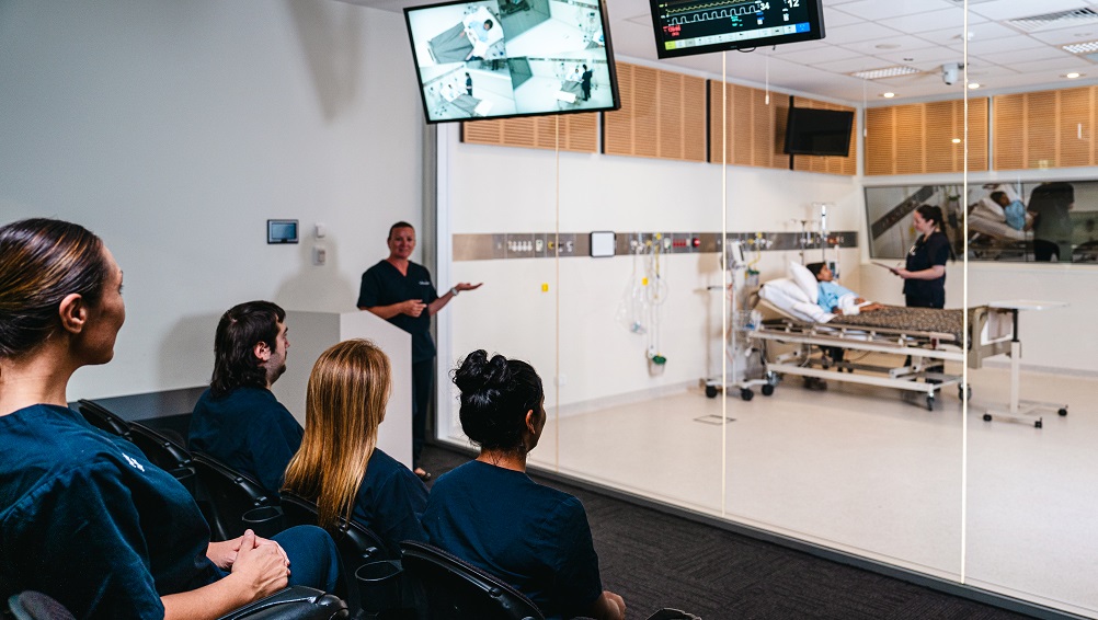 Student listen to teachers as they watch a simulated exercise in a training ward.