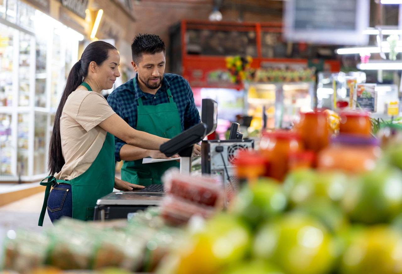 Cashier training a Latin American man on how to use the cash register at a supermarket